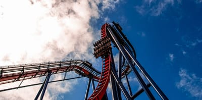 A red steel dive roller coaster photographed from directly below, its train of riders held at the top of a vertical drop against a blue sky.