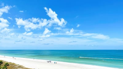 A wide white-sand beach on the Gulf of Mexico photographed from several storeys up, with turquoise shallows over a sandbar, a vegetated dune and cabbage palms at the lower left, and a few shade canopies on the sand.