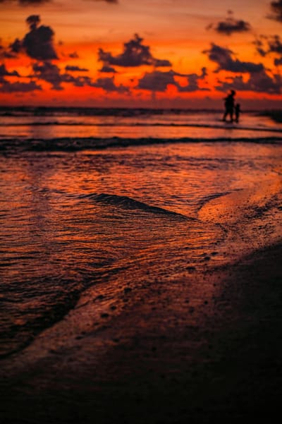 Two figures silhouetted at the waterline of a Gulf beach beneath an orange and red sunset sky.