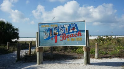 A Sarasota County sign at a beach access path reading Siesta, with white sand and the Gulf behind it.