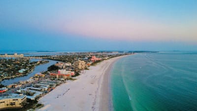 Aerial view at dusk looking south along St. Pete Beach, Florida, showing Gulf Boulevard hotels on the left, a wide white sand beach in the centre, the pink Don CeSar hotel in the middle distance, and the turquoise Gulf of Mexico on the right.