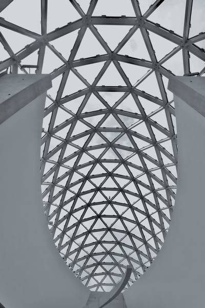 Looking upward through a pointed oval opening in a concrete ceiling at a curved steel-and-glass lattice vault under an overcast sky.